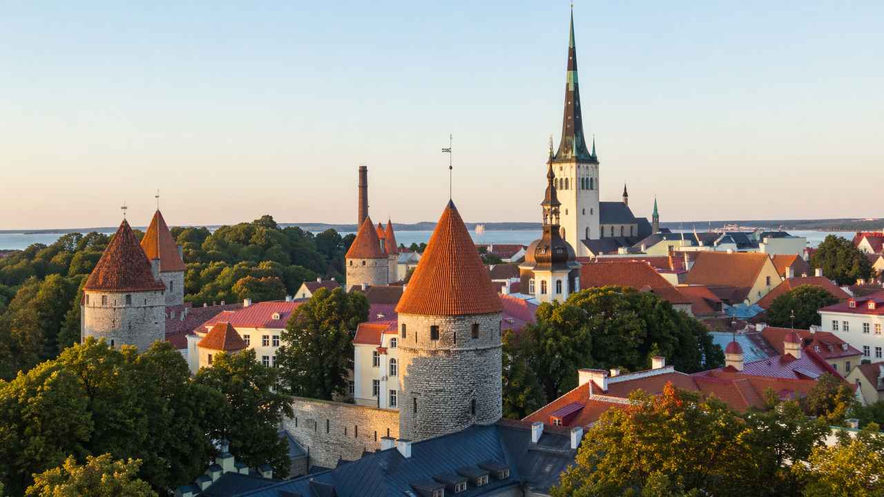Panoramic view of Tallinn Old Town in Estonia at golden hour, with medieval stone towers, red rooftops, church spires, and the sea in the distance.