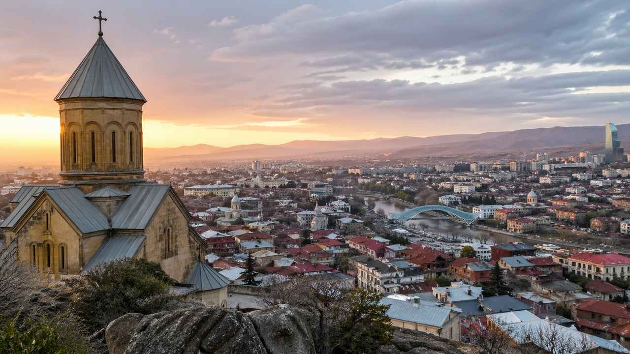 Stone church overlooking Tbilisi, Georgia, at sunset, with the city, river, bridge, and distant hills under a dramatic evening sky.
